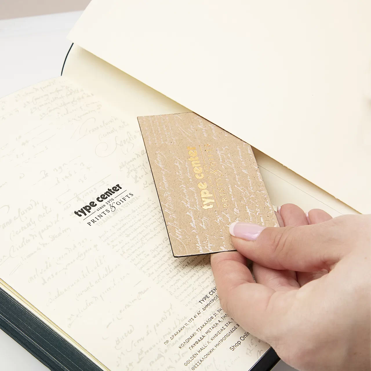 ανοιχτό μπεζ ημερολόγιο με ένα χέρι να κρατάει μια σκούρη μπεζ κάρτα με χρυσά γράμματα