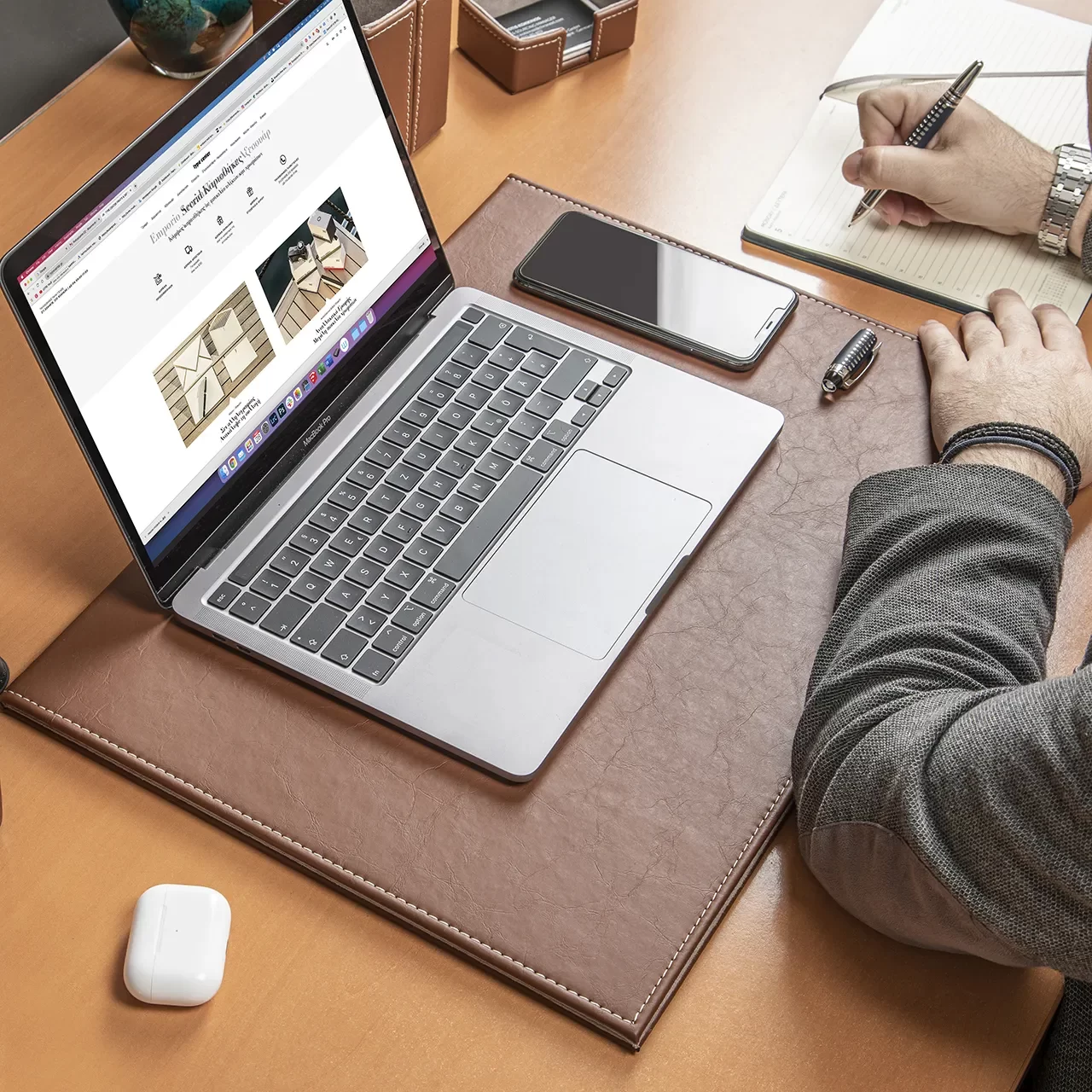 a close up of a wooden desk that on top of it has an open silver laptop that under it has a brown leather deskmate also the desk it has an airpods case a smartphone also there is hands that rest on the desk and also write on a notebook