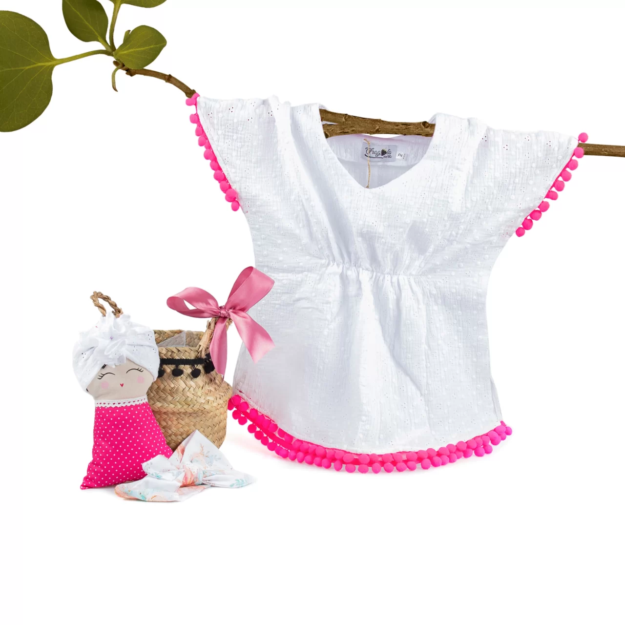 a white baby shirt with pink details on the seams that hangs from a tree branch and beside it it has a cloth doll and a small rope basket with a pink bow